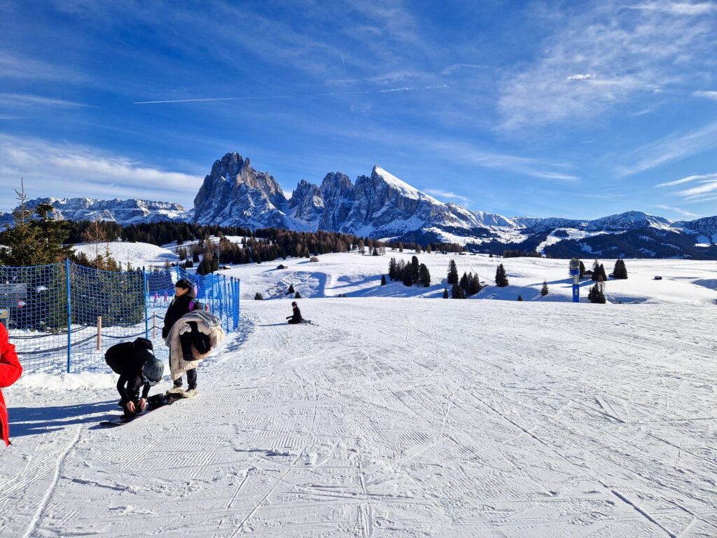 A wide groomed ski slope opens onto rolling snow covered meadows and evergreen trees, with dramatic jagged mountain peaks rising in the distance. A few skiers pause near blue safety fencing while others prepare their gear, all set against a bright blue winter sky.