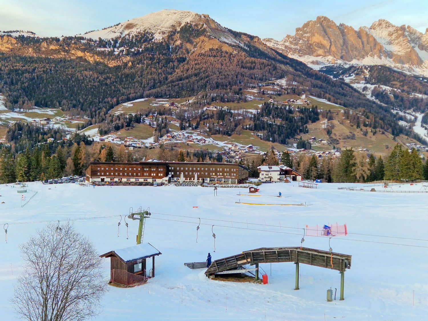 A snowy ski area sits above an alpine village with lift lines and beginner terrain in the foreground. Forested hills rise behind scattered chalets and farms with rugged mountain peaks catching warm light in the distance.
