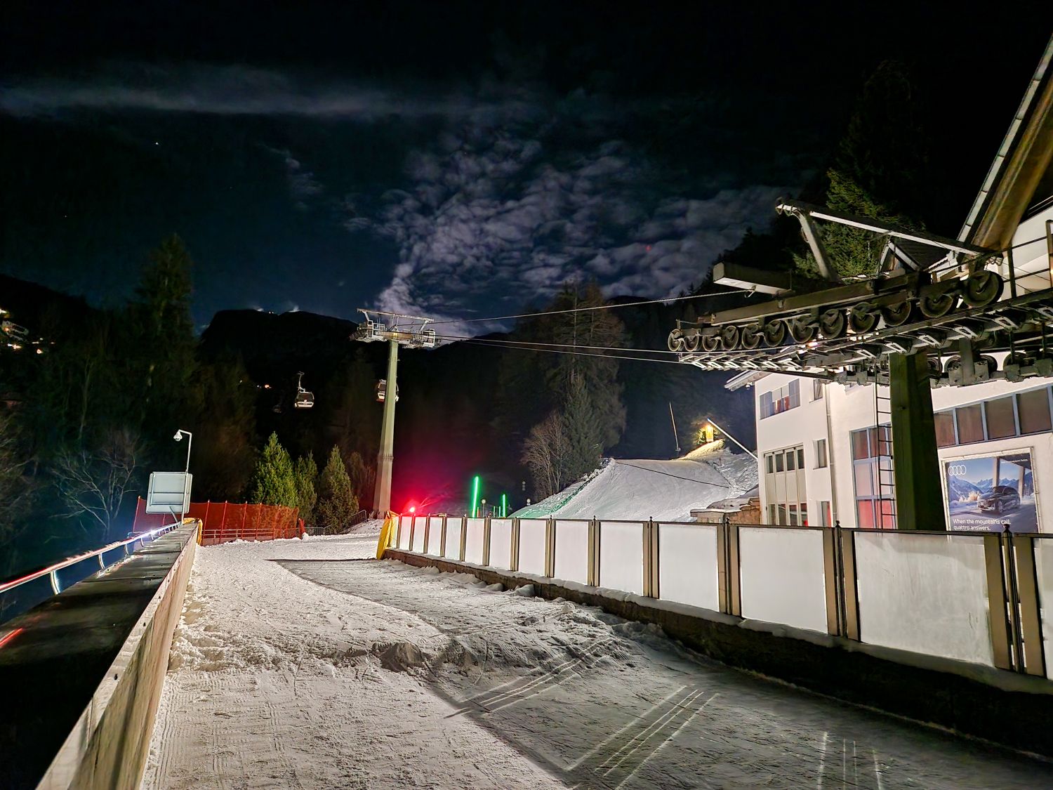 A snowy ski lift area is lit at night with chairlift cables overhead and a quiet run leading away from the station. Artificial lights illuminate the snow and nearby trees under a dark sky with textured clouds creating a calm after hours alpine atmosphere.