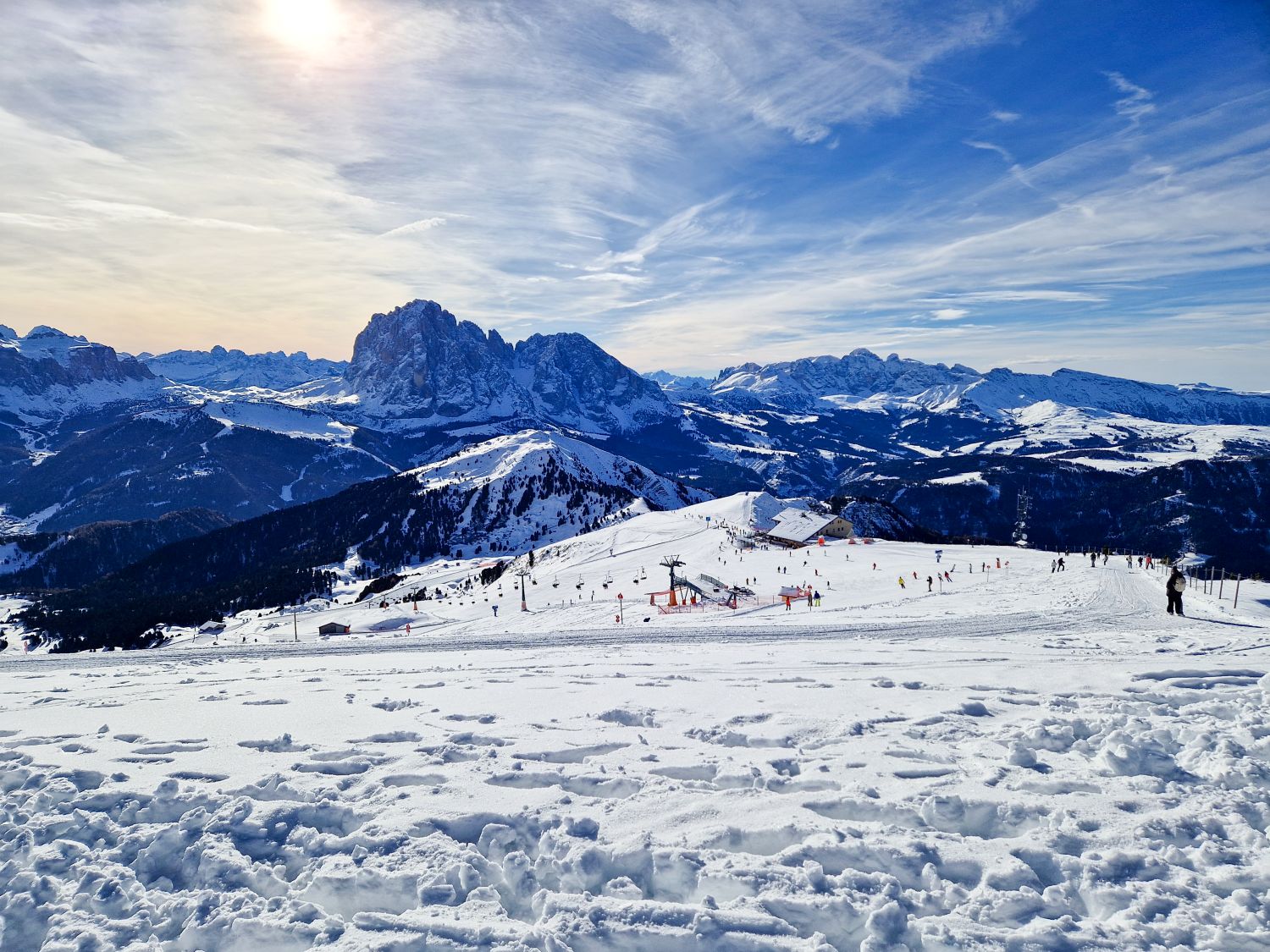 A panoramic view looks across a high alpine ski area with wide snow covered slopes and scattered skiers below. Layered mountain ridges and dramatic rocky peaks stretch across the horizon under a softly glowing winter sky, emphasizing the vast open landscape.