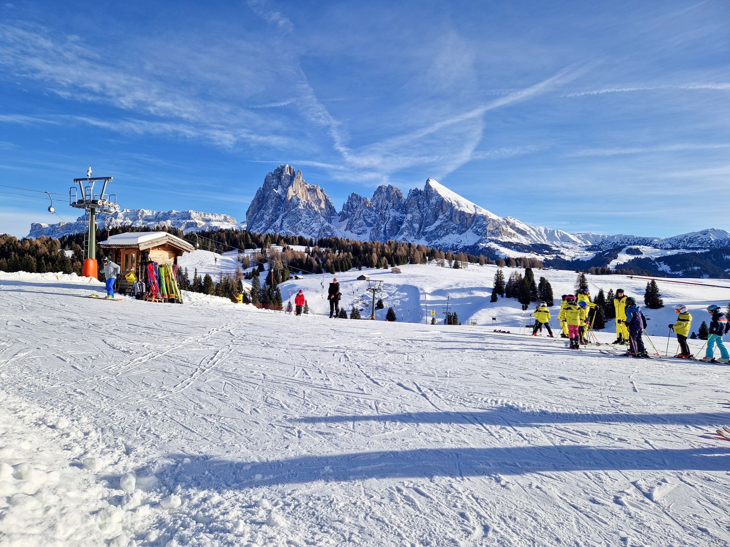 A wide beginner ski area opens onto a panoramic alpine landscape, with children in bright ski gear gathered for a lesson on a gentle slope. Snow covered meadows and evergreen trees stretch toward dramatic jagged mountain peaks under a vivid blue winter sky.