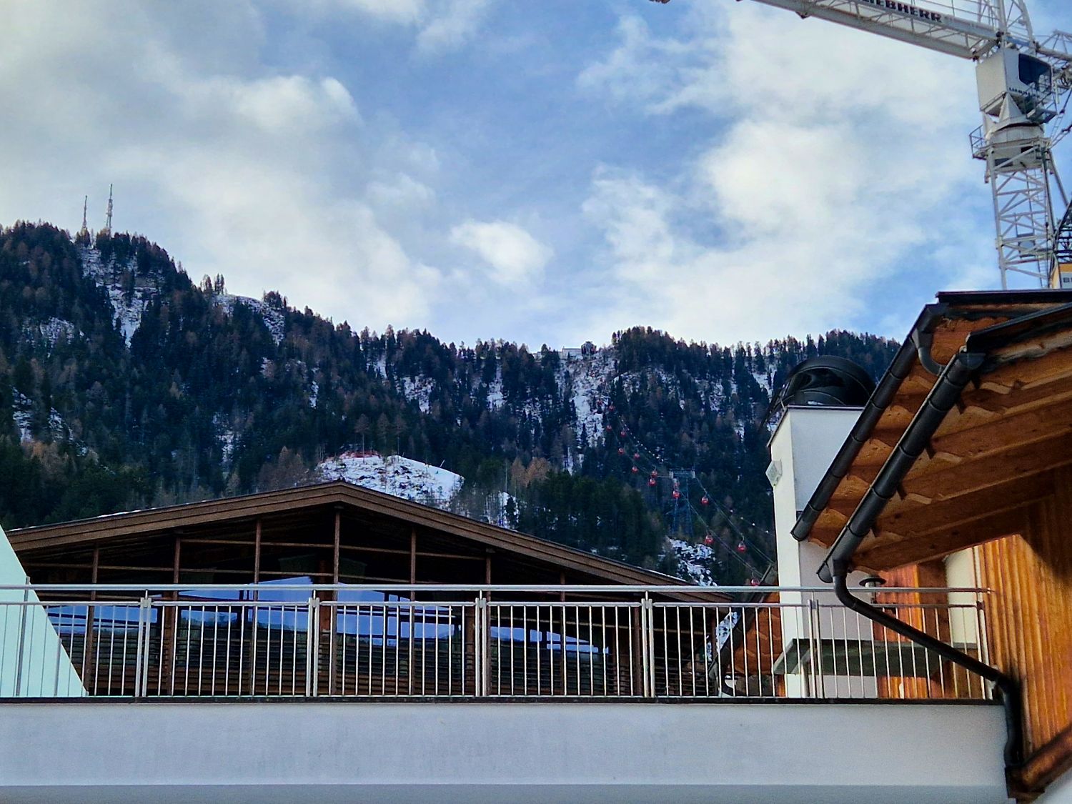 A view of snow-dusted mountains behind wooden buildings and a metal railing, with a construction crane and a ski lift line visible against a partly cloudy sky.