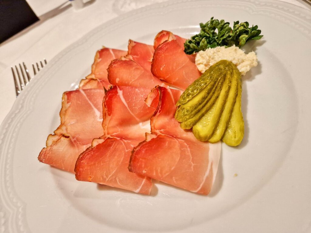 Thin slices of cured meat arranged on a white plate, served with sliced pickles, a dollop of horseradish, and a small garnish of fresh greens.