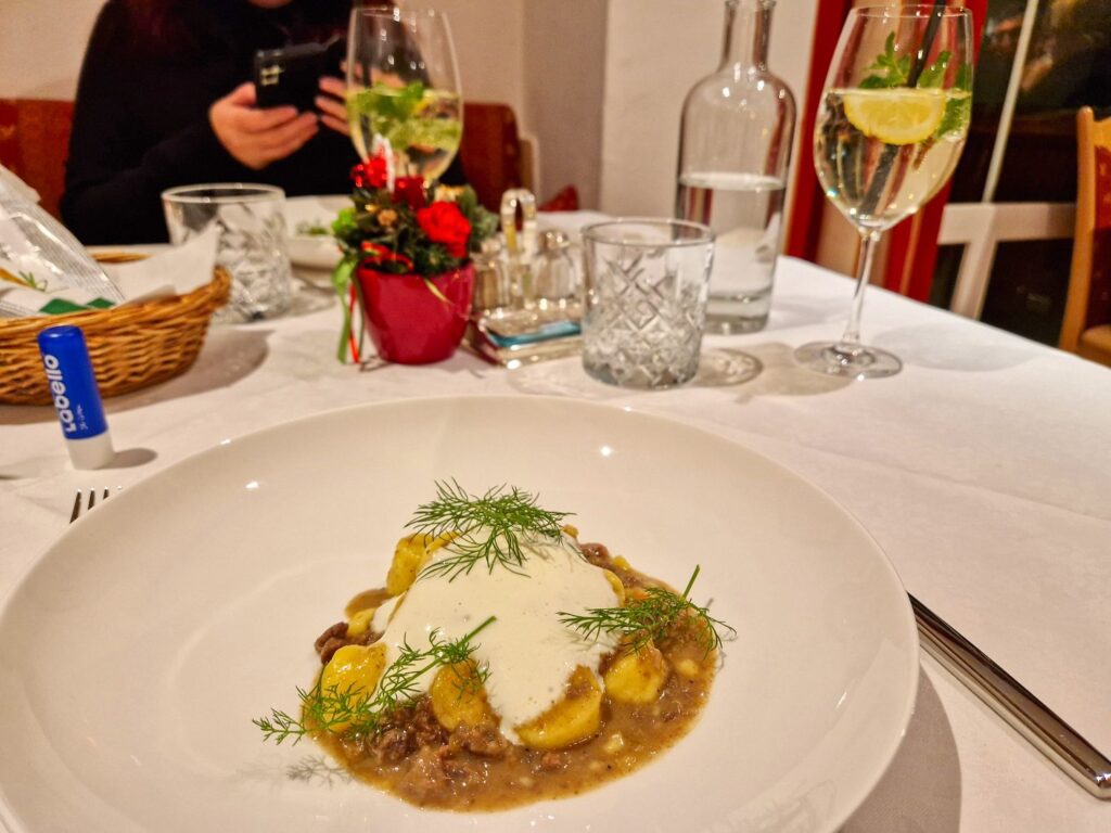 A plate of gnocchi with creamy sauce and herbs on a white tablecloth, with wine, water, and a small flower arrangement. A person in the background is using a phone.