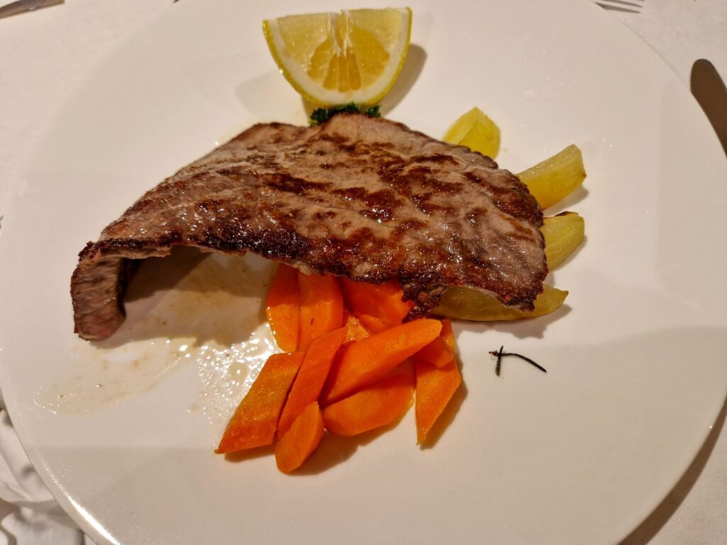 A grilled steak served on a white plate with sliced cooked carrots, pieces of yellowish vegetables, a lemon wedge, and a small garnish of parsley.
