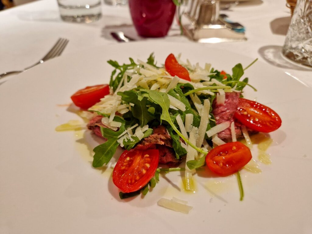 A plate of salad with sliced steak, arugula, halved cherry tomatoes, and grated cheese, drizzled with dressing, on a white tablecloth with glassware and cutlery in the background.