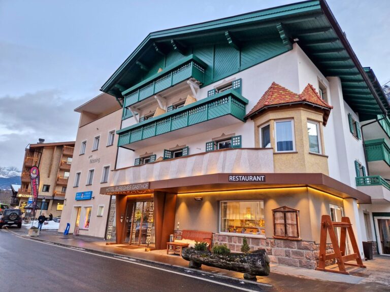 A street view of a multi-story Alpine-style hotel and restaurant with green balconies, large windows, and a wooden bench outside, set against a cloudy sky and mountain backdrop.