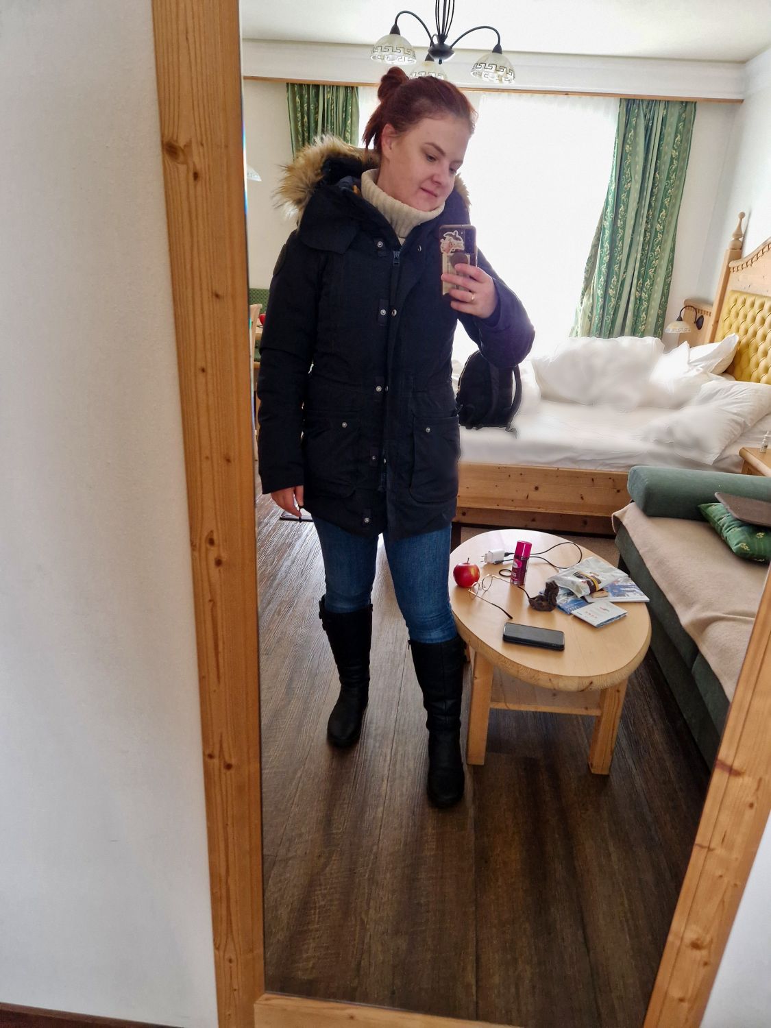 A woman in a dark winter coat, jeans, and boots takes a mirror selfie in a cozy room with a wooden floor, a small round table, and a couch with a blanket in the background.