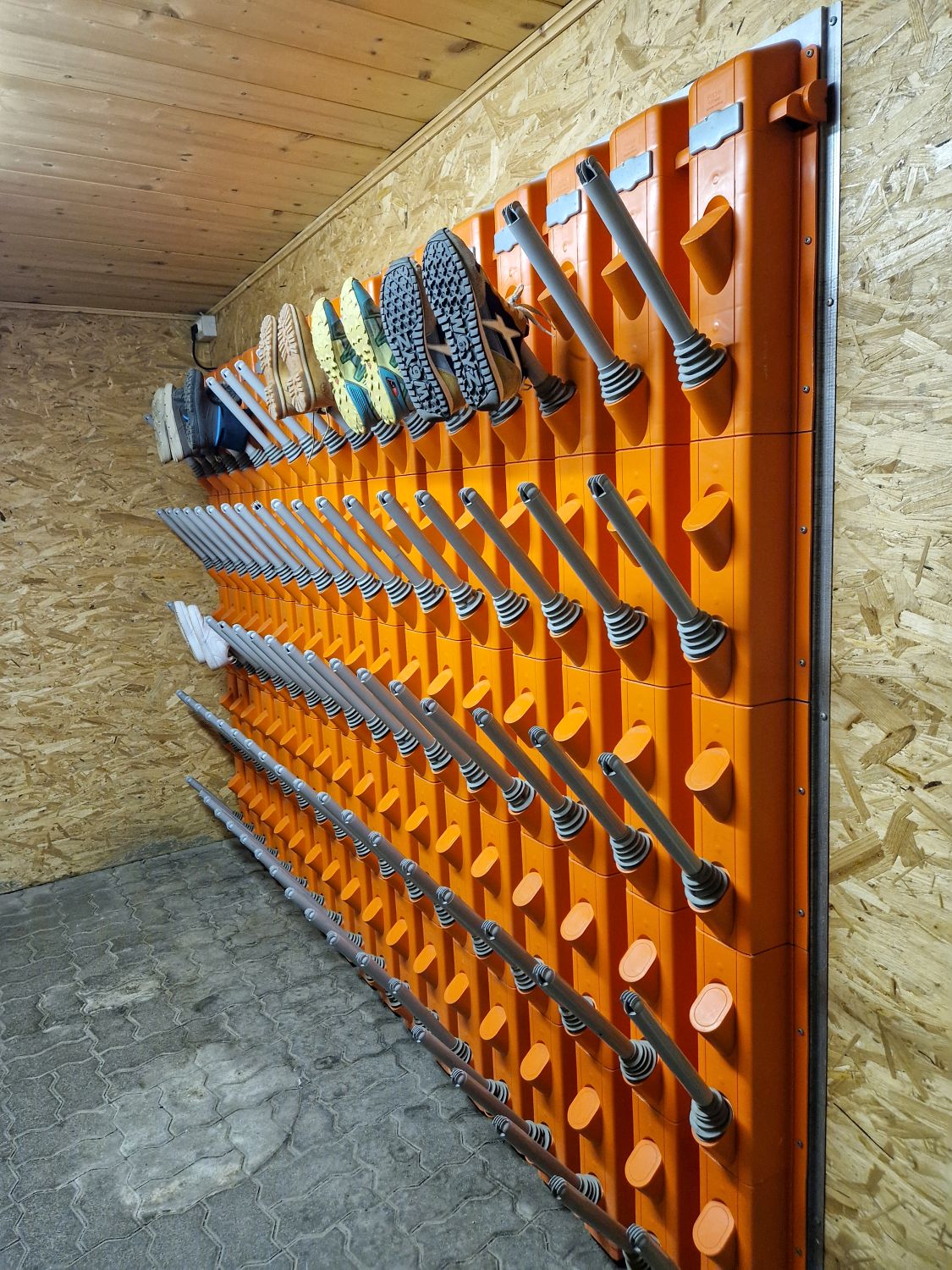 Rows of orange boot dryers are mounted on a wooden wall in a room, with several pairs of boots placed upside down on the drying tubes. The floor is tiled, and the ceiling and walls are made of wood panels.
