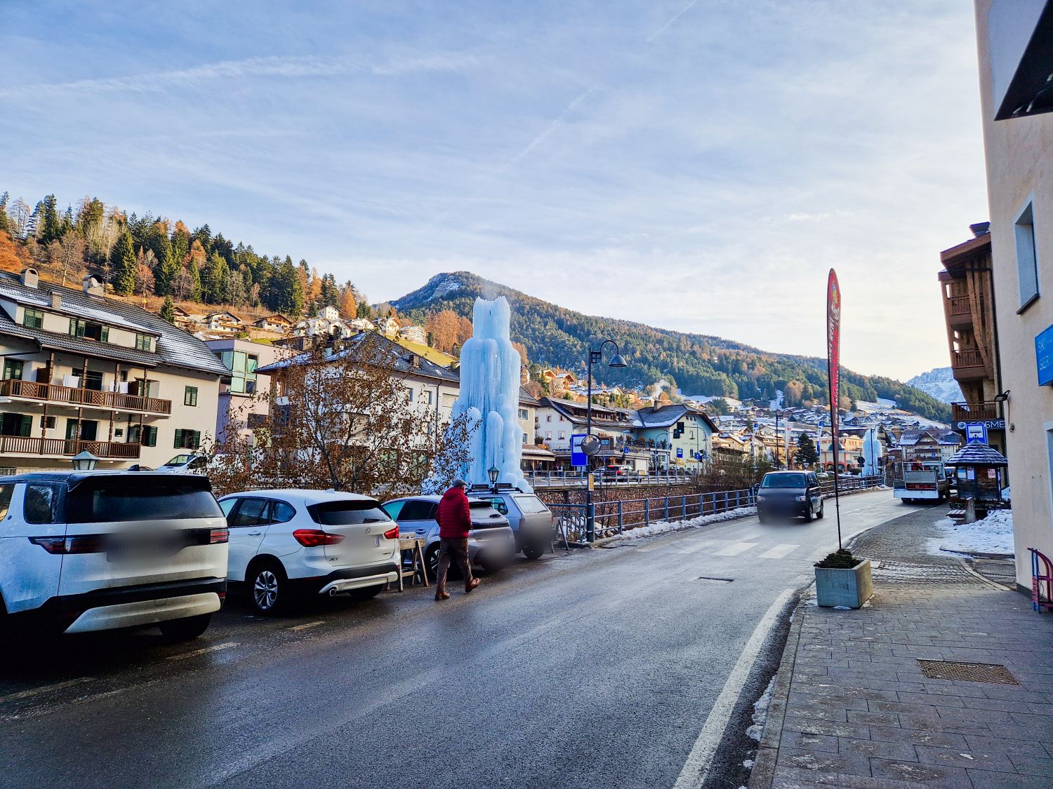 A street in a small mountain town with parked cars, a person walking, and a tall frozen ice sculpture. Hills with autumn trees and scattered houses are visible in the background under a clear sky.