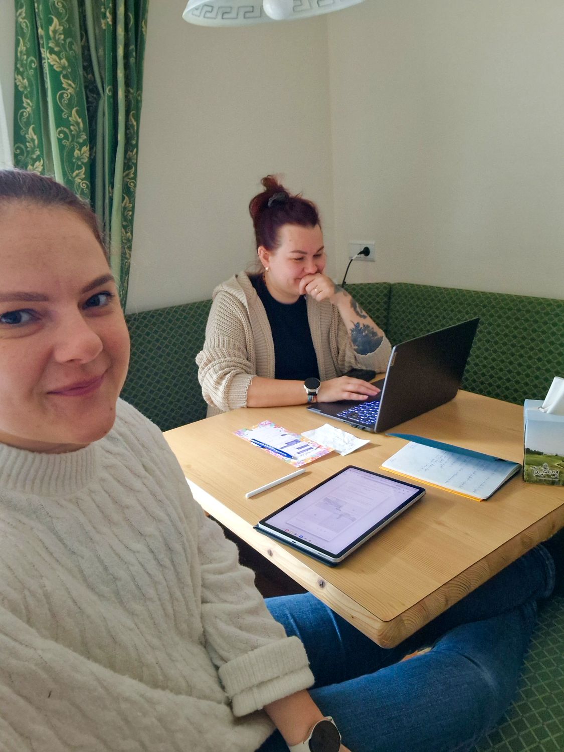 Two women sit at a wooden table with a laptop, tablet, and papers. One is smiling at the camera while the other works and laughs. The setting appears to be a cozy, well-lit room with green patterned curtains.