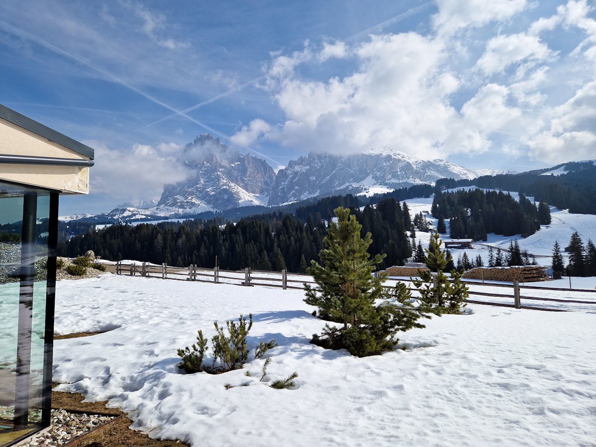 Snow covered alpine landscape with small pine trees in the foreground and a wooden fence stretching across a snowy hillside. In the distance rugged mountains rise through drifting clouds under a bright blue sky, viewed from the edge of a modern house.