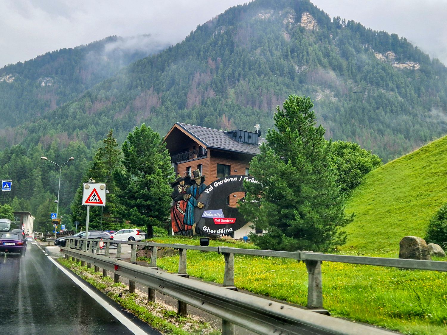 Roadside view of a mountain village entrance with a Val Gardena sign, evergreen trees, and misty forested mountains rising behind a chalet-style building. Cars drive along a wet road, emphasizing arrival into an alpine valley.
