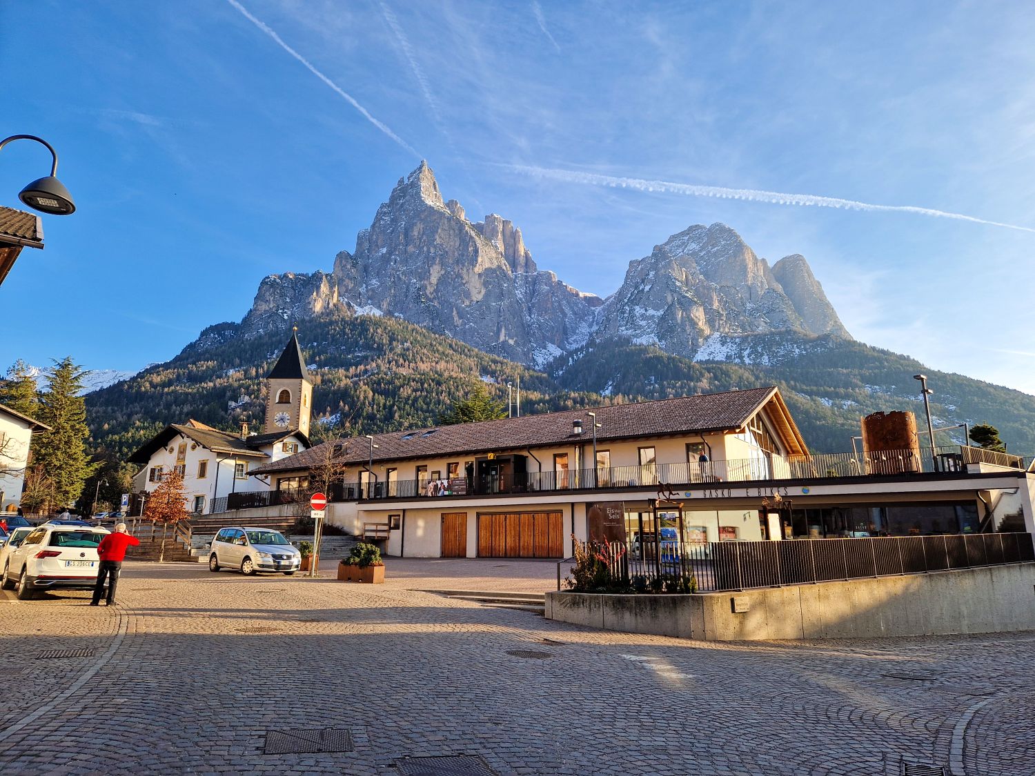 A quiet village square with a church tower and low buildings sits beneath dramatic jagged mountain peaks dusted with snow under a bright blue sky. A person in a red jacket stands near parked cars, adding scale to the towering alpine landscape behind the town.