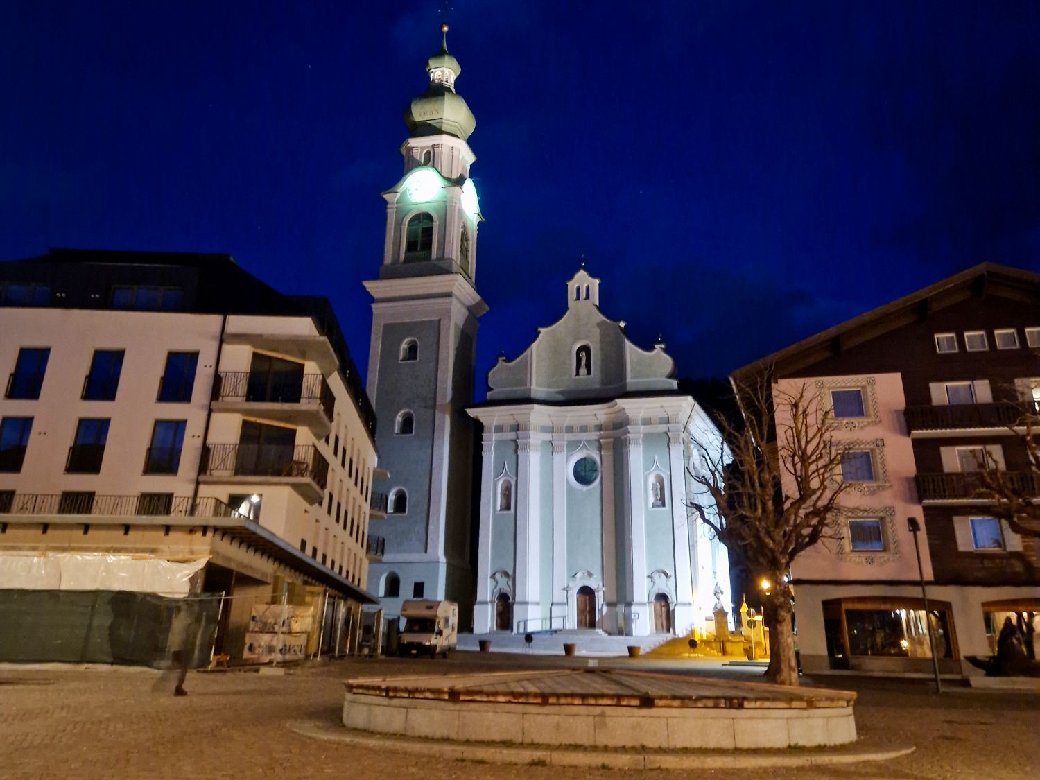 Nighttime view of a church with a tall bell tower and onion dome in Dobbiaco in the Dolomites illuminated against a deep blue sky in a quiet town square. The pale facade of the church stands between modern alpine buildings with bare trees and empty pavement, creating a calm evening atmosphere.