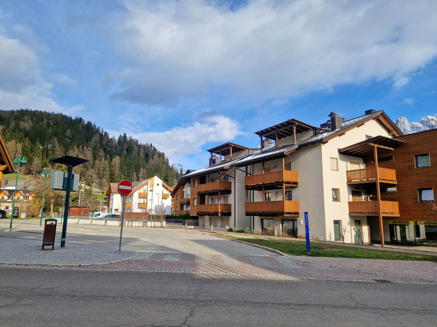 Modern alpine apartment buildings with wooden balconies line a quiet street in a mountain village, with forested hills rising behind the homes. A pedestrian crossing and street signs sit in the foreground under a partly cloudy blue sky, giving a calm residential feel.