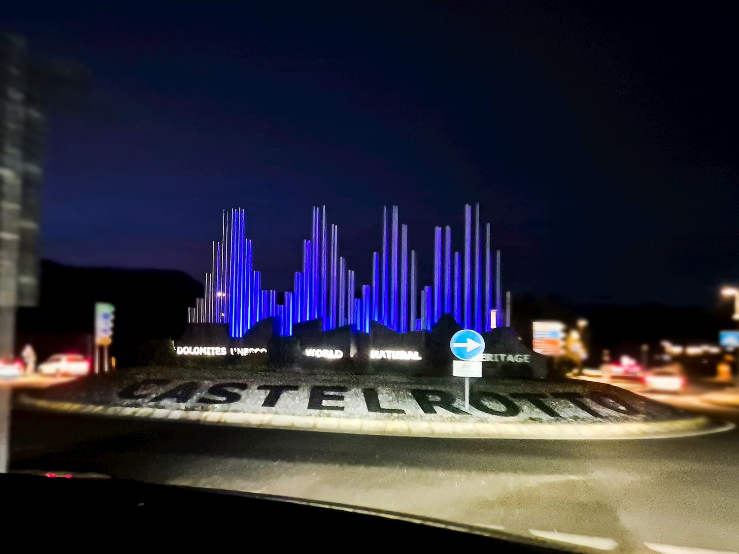 Alt text. Nighttime view of a roundabout in Castelrotto with an illuminated art installation shaped like mountain peaks glowing blue against a dark sky. The base of the display reads Dolomites UNESCO World Natural Heritage while traffic signs and blurred car lights surround the roundabout, emphasizing arrival in the Dolomites.