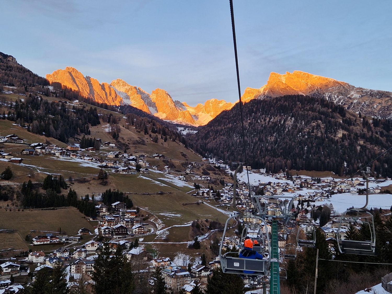 Winter view in Santa Cristina showing a chairlift ascending over snowy slopes with glowing pink alpenglow on the Dolomite peaks during enrosadira. Soft pastel light reflects off the mountains at sunset, creating a calm alpine winter atmosphere.