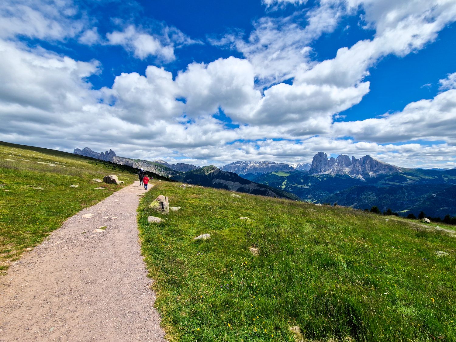 A wide gravel hiking path winds through a green alpine meadow with two hikers walking toward distant jagged mountain peaks. Rolling hills, rocky outcrops, and dramatic cliffs stretch across the horizon under a bright blue sky filled with large white clouds. The image conveys an open scenic mountain landscape and a peaceful hiking experience in nature.