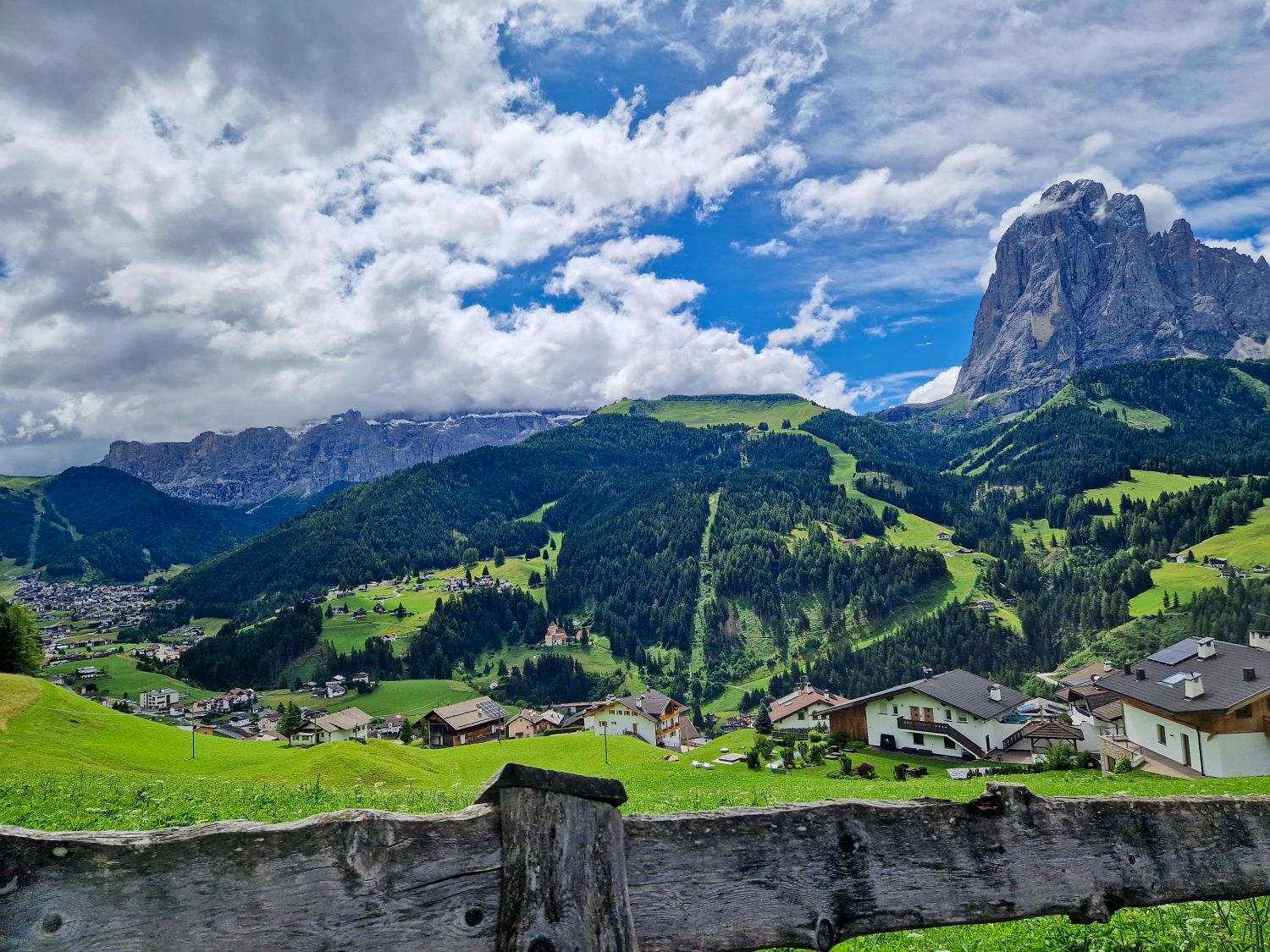 Mountain trail view near Santa Cristina with the Sassolungo massif towering above a paved path leading toward Baita Seurasas. Green alpine meadows and scattered trees frame the scene, emphasizing the scale of the rocky Dolomite peaks on a clear summer day.