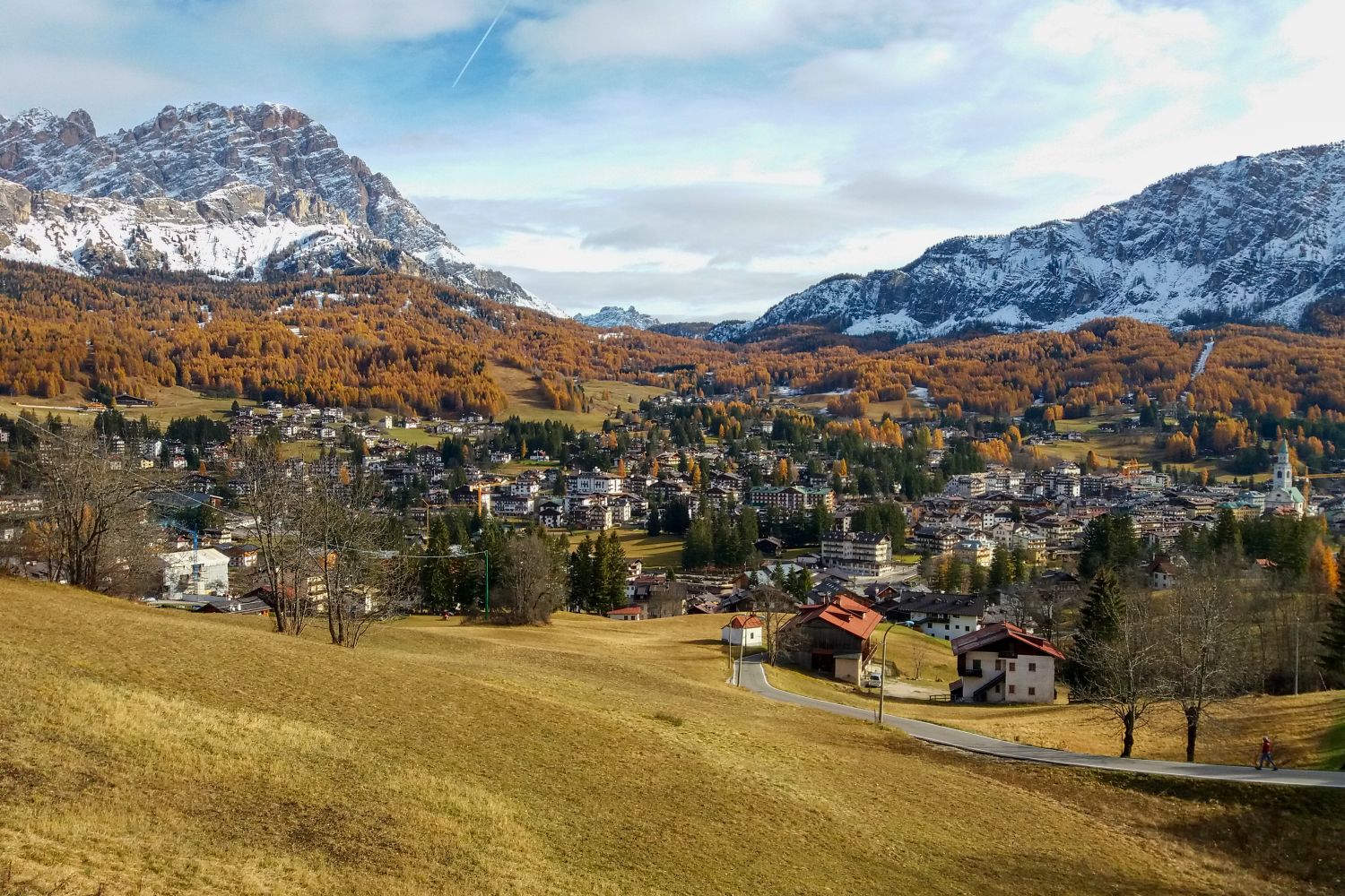 Things to Do in the Dolomites That Are Actually Worth It 41 Wide autumn view of a town nestled in a mountain valley, with golden trees covering the hillsides and snow-dusted peaks in the distance. Scattered houses and church towers sit among fields and winding roads.