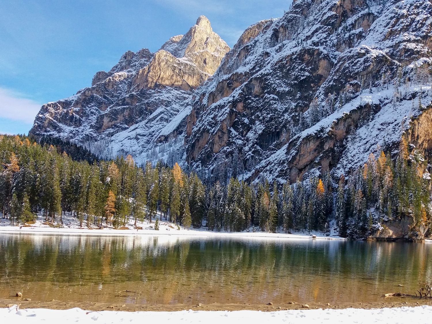 Clear alpine lake reflecting a dramatic rocky mountain face partially covered in snow, with dense evergreen trees lining the shore. The calm water mirrors the cliffs and forest, creating a serene mountain scene.