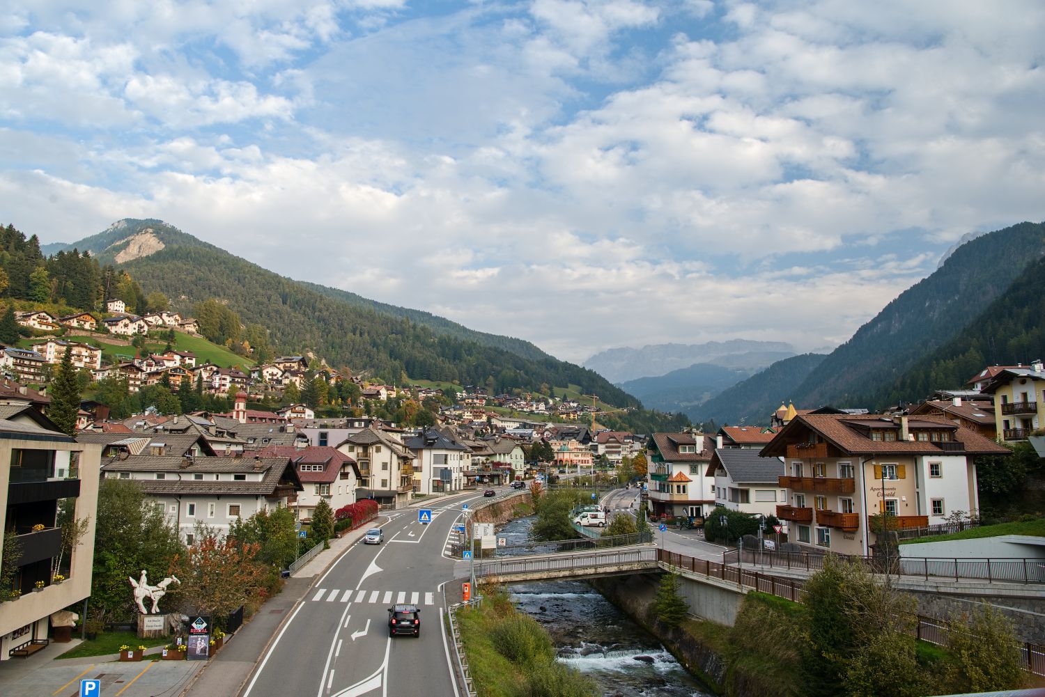 Alpine village spread across a valley with clusters of houses and winding roads beside a narrow river. Forested mountains rise on both sides under a sky filled with scattered clouds.