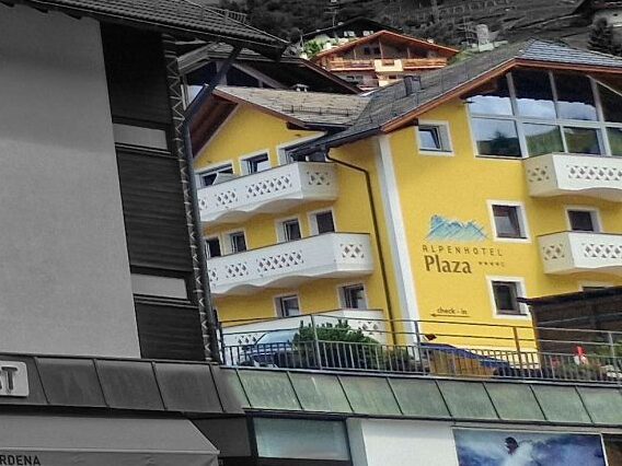 Street corner with a modern hotel building labeled "DOSSES LIVING THE DOLOMITES" above an "INTERSPORT" shop. Surrounding buildings and hills are mostly in black and white while a yellow hotel stands out in color on the right.