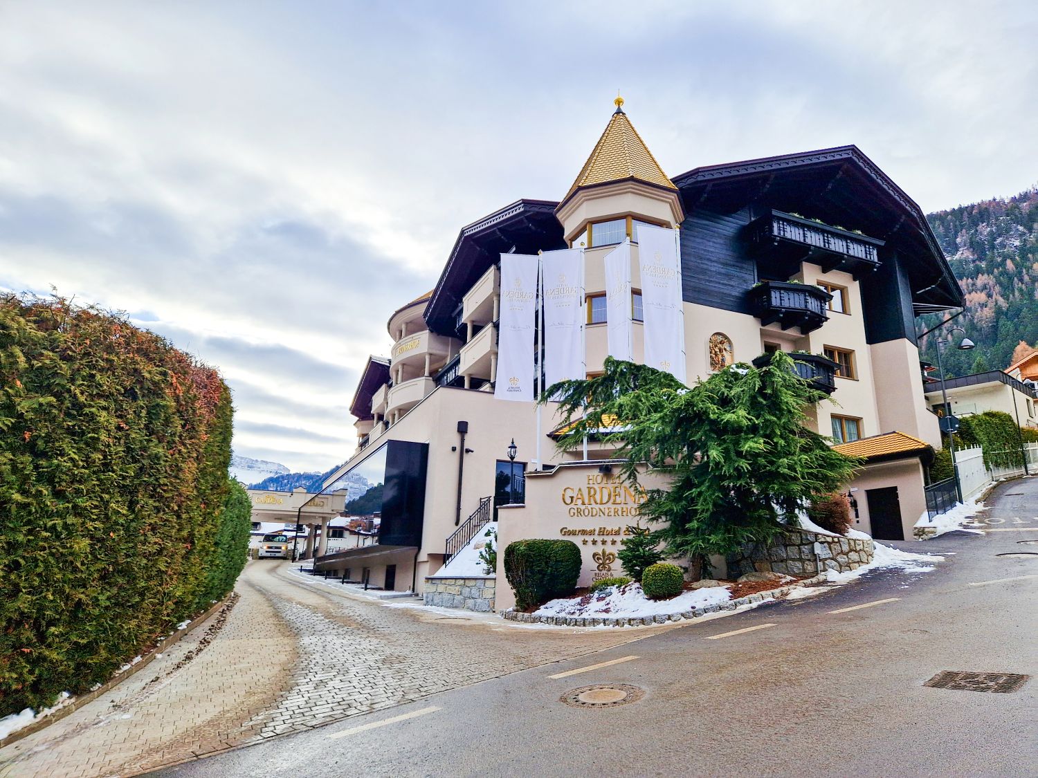 arge alpine hotel with a tower and banners displaying "GARDENA GRODNERHOF" near the entrance. A curved driveway leads past trimmed bushes and patches of snow with mountains in the background.