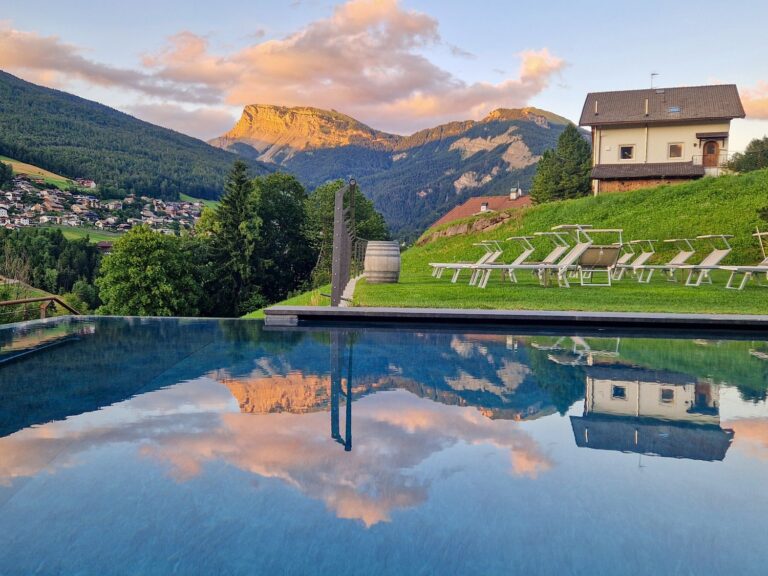 Infinity pool at Digon hotel in Val Gardena reflecting a mountain landscape at sunset with warm light hitting the rocky peaks. Lounge chairs and a small house sit on a grassy terrace overlooking a valley filled with clustered homes and trees.