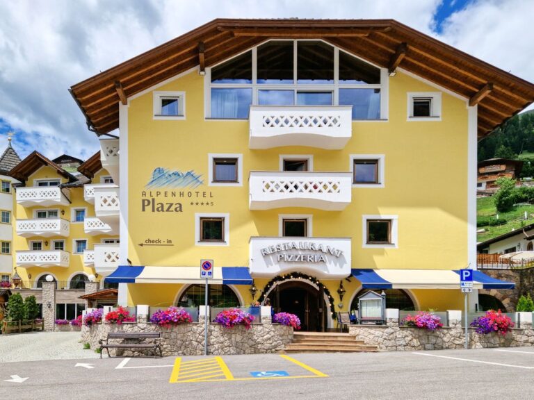 Alpenhotel Plaza in Val Gardena with pale yellow walls, white balconies, and a restaurant entrance lined with colorful flower boxes, viewed from across a parking area. Green alpine hills and traditional mountain buildings surround the hotel under a dramatic cloudy sky, showcasing one of the hotels in Val Gardena.