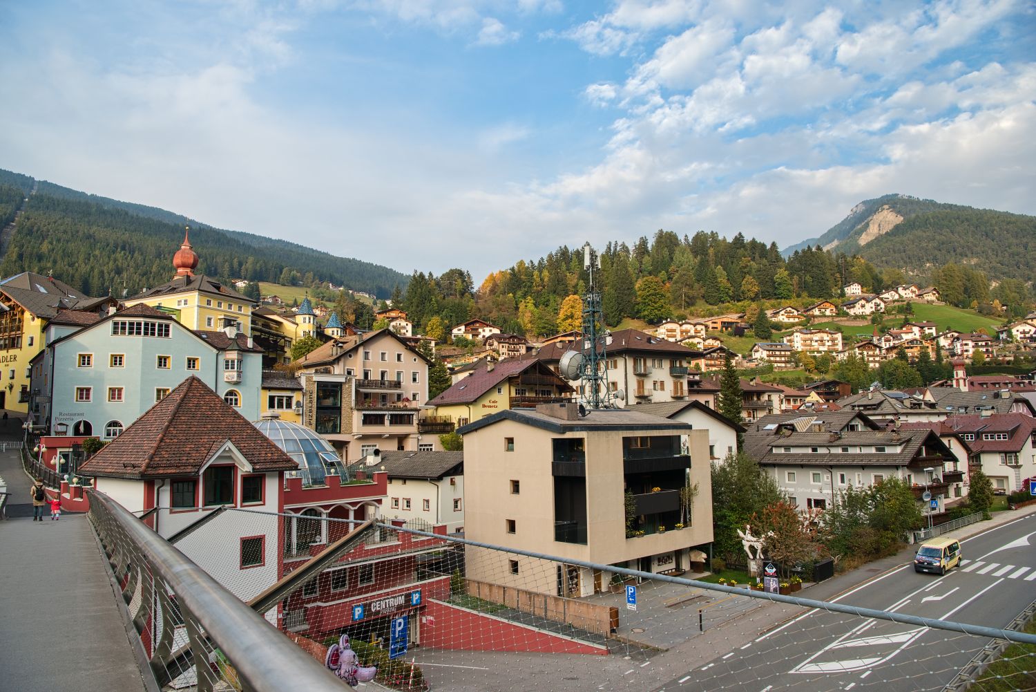 View from a pedestrian bridge overlooking an Ortisei with pastel buildings and sloped roofs clustered along the hillside. A road with light traffic runs below, and forested mountains rise in the background under a partly cloudy sky.