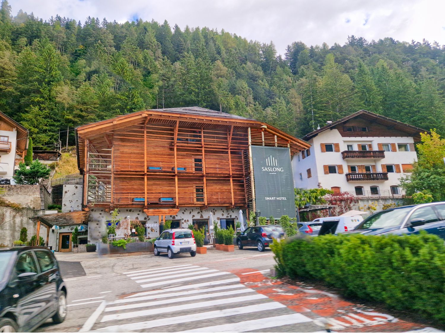 Wooden alpine style hotel with slatted exterior and balconies sits at the base of a forest covered hillside. A vertical sign on the building reads "SASLONG" and "SMART HOTEL" while cars are parked in front near a crosswalk.
