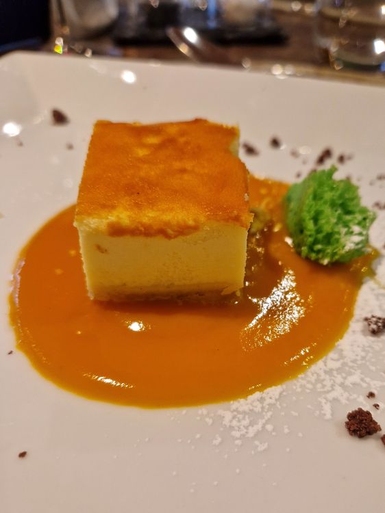 A square slice of cheesecake sits in a pool of glossy orange sauce on a white dessert plate dusted with powdered sugar. A small green garnish and scattered chocolate crumbs decorate the plate beside the golden-topped dessert.