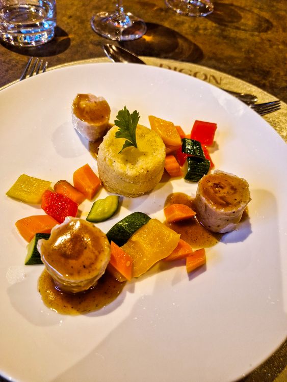 Seared medallions of meat are arranged around a creamy molded side dish and colorful diced vegetables on a white fine dining plate. Brown sauce is drizzled beneath the meat while parsley garnish adds a fresh touch to the elegant presentation.