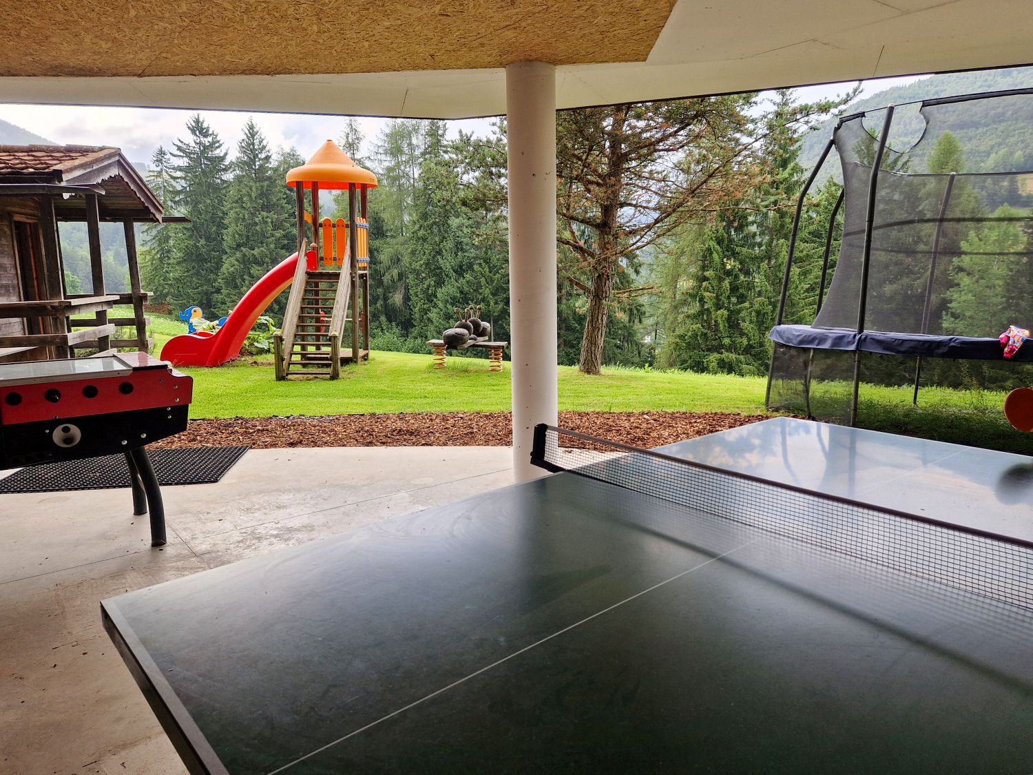 A covered outdoor play area features a ping pong table, foosball table, trampoline, and colorful playground slide surrounded by tall pine trees. The grassy yard overlooks a forested mountain landscape, creating a quiet family-friendly recreation space.
