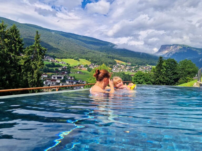 Kristine and Emma relax in an infinity pool overlooking a green mountain valley called Val Gardena dotted with small villages and pine trees. Cloud-covered peaks, including Seceda, rise in the background while sunlight reflects across the calm blue water.
