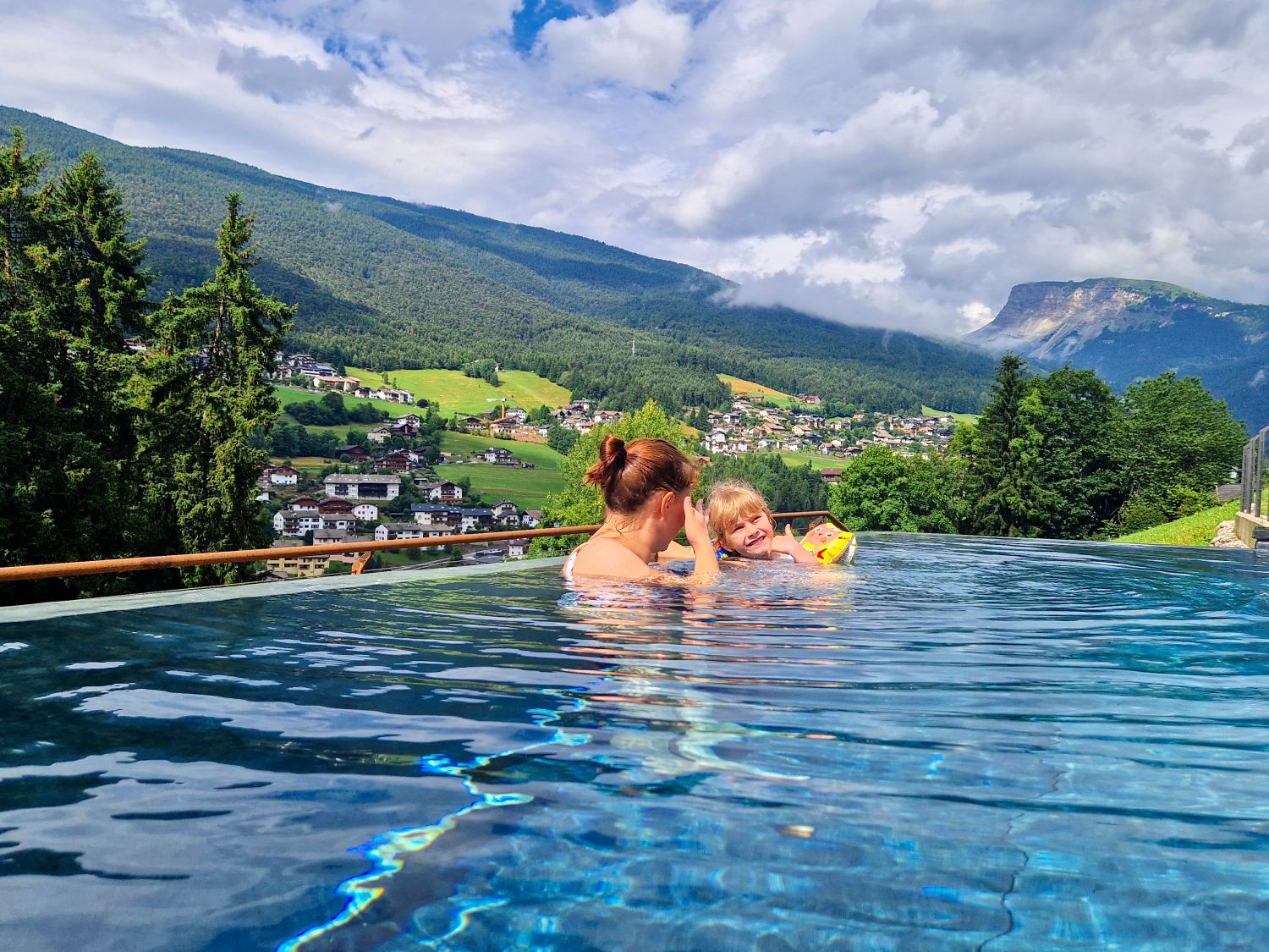 Kristine and Emma relax in an infinity pool overlooking a green mountain valley called Val Gardena dotted with small villages and pine trees. Cloud-covered peaks, including Seceda, rise in the background while sunlight reflects across the calm blue water.