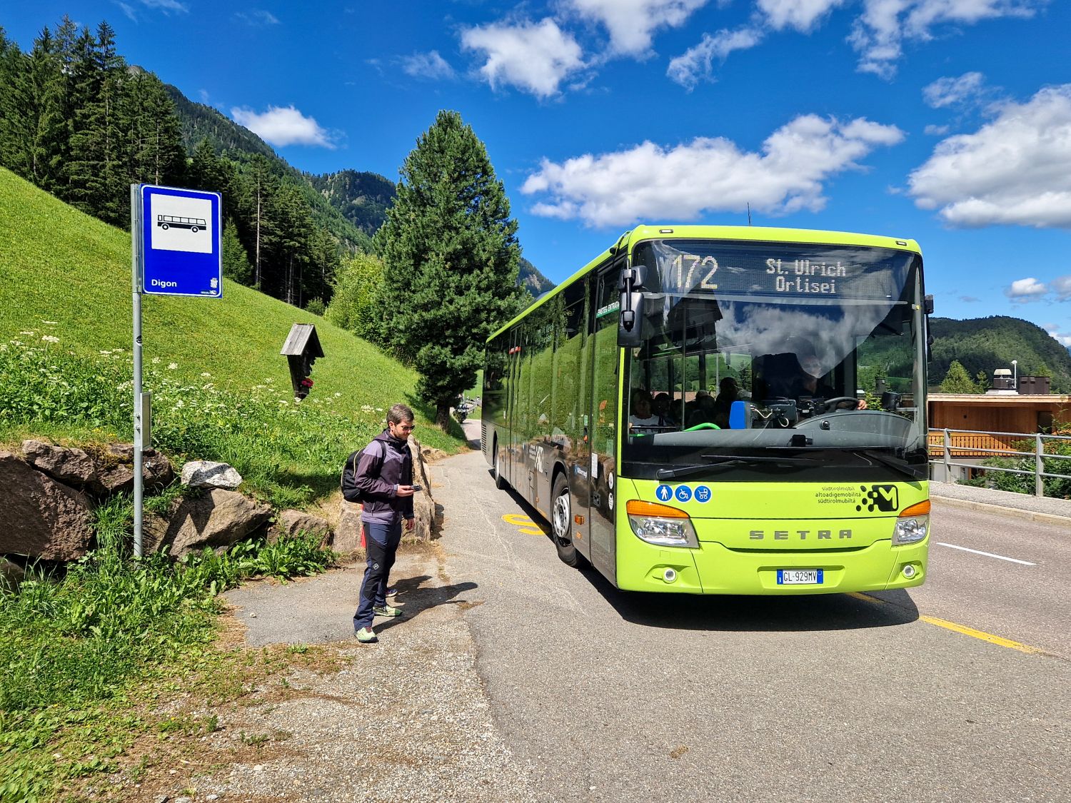 A bright green public bus marked “172 St. Ulrich Ortisei” stops beside a rural roadside bus sign in a mountain valley. A man with a backpack waits nearby while green hills, pine trees, and a blue sky with scattered clouds surround the scene.