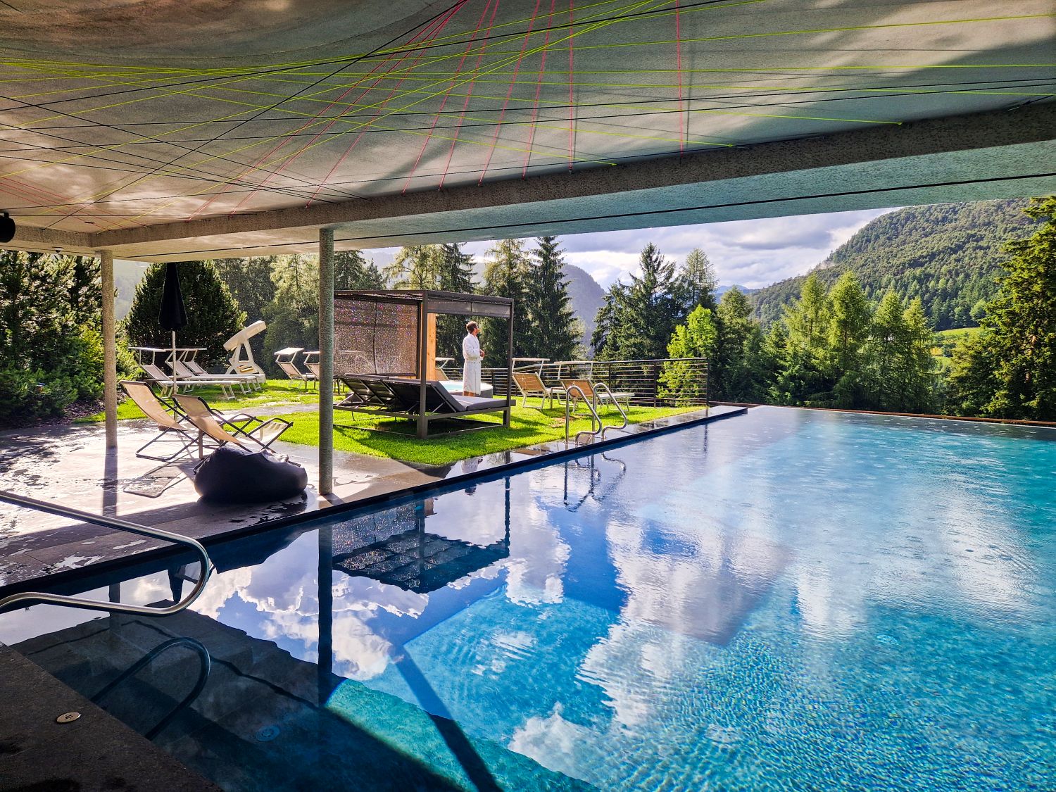 A covered outdoor pool at Hotel Digon reflects clouds and surrounding pine forests while lounge chairs and shaded daybeds sit beside the water. Sunlight brightens the mountain spa setting as a person in a white robe - Michal - stands near the edge of the pool overlooking the valley.