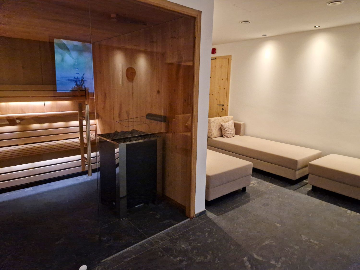 A modern spa relaxation area in hotel Digon features a glass-front sauna with warm wooden benches and soft ambient lighting. Beige lounge chairs line the quiet room beside dark stone flooring and light wood accents for a calm wellness atmosphere.
