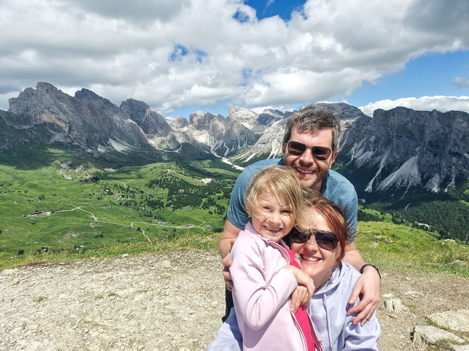 Family of three smiling together on a mountain viewpoint with dramatic rocky peaks, green valleys, and winding trails stretching across the landscape behind them. An adult man and woman wearing sunglasses embrace a young child in a pink jacket as they pose against a backdrop of rugged mountains and large white clouds in a blue sky.