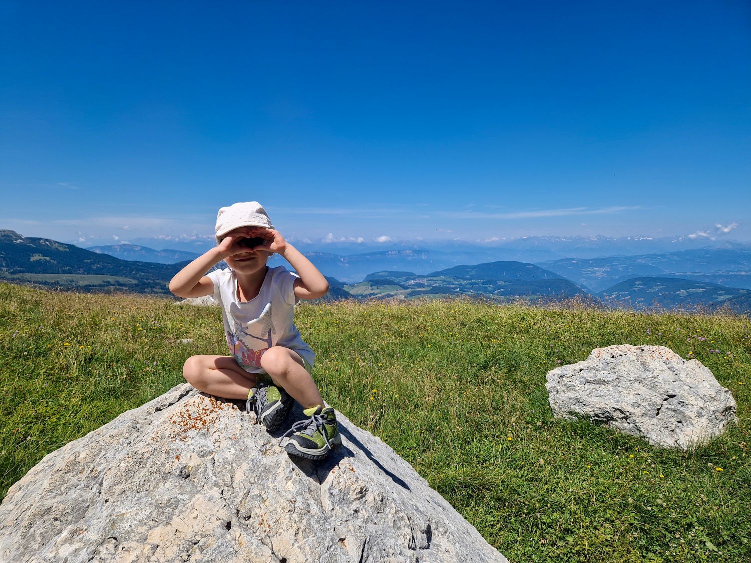 Emma sits on a large rock along the Latemar Panorama Trail near Obereggen, shading her eyes as she looks out across rolling alpine meadows and distant mountain peaks. The clear blue sky and wide-open views capture the peaceful scenery of this family-friendly hike in the Dolomites.