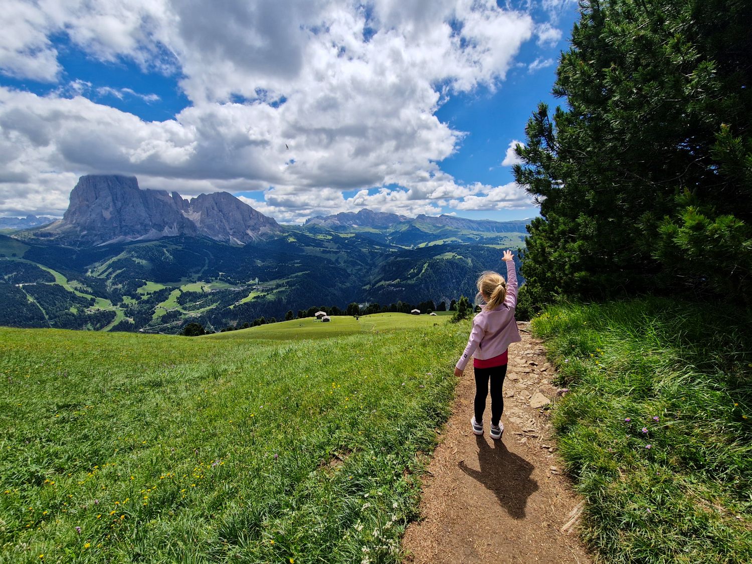 Emma hikes toward Mt. Pic above Santa Cristina in Val Gardena wearing hiking sandals on a narrow trail through a green alpine meadow. The rolling hills and rugged Dolomite peaks illustrate the varied terrain where footwear with good sole grip, including sturdy hiking sandals, can be suitable for many routes in the Dolomites.