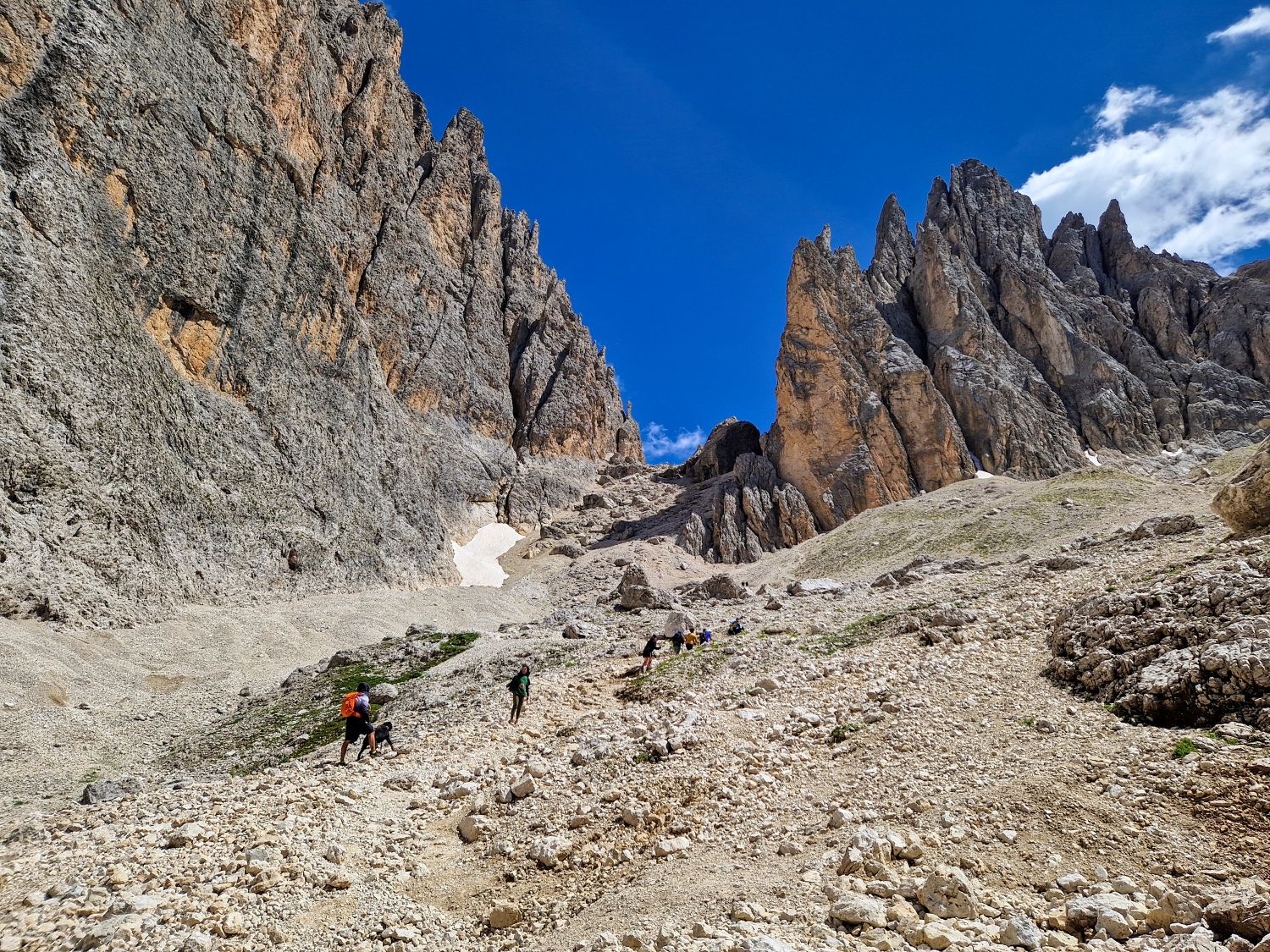 Hikers make their way across the rocky scree slopes of the Sassolungo Loop hike in Val Gardena, surrounded by towering limestone cliffs and jagged Dolomite peaks. The rugged alpine landscape stretches beneath a deep blue sky, showcasing one of the region's most dramatic hiking routes.
