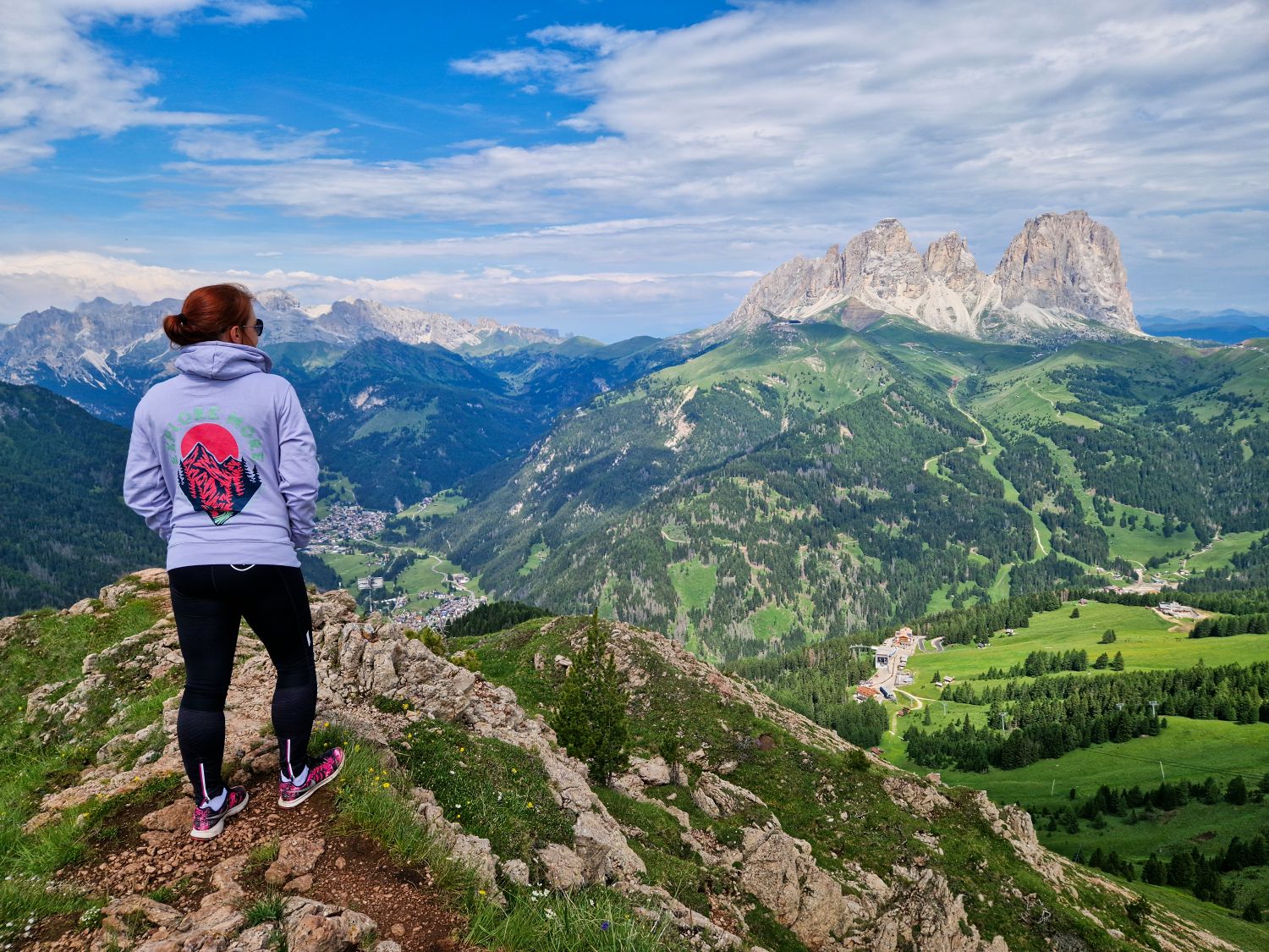A hiker stands on a rocky viewpoint overlooking the green valleys and dramatic peaks of the Dolomites near Val Gardena. The rugged mountain terrain highlights the type of landscape where the best hiking shoes for the Dolomites provide comfort, grip, and stability on uneven trails.