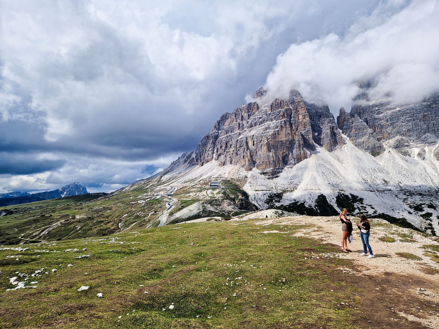 Two visitors stand on the Cadini di Misurina viewpoint trail overlooking dramatic limestone peaks, with Tre Cime di Lavaredo partially hidden by low clouds in the background. Rifugio Auronzo is visible below among the rocky slopes, while dark storm clouds gather over the surrounding Dolomite landscape.